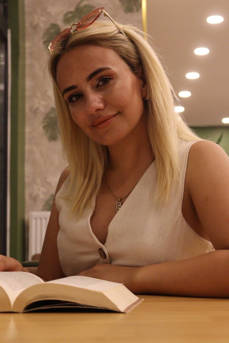 A young woman with blonde hair reads a book indoors under warm lighting.