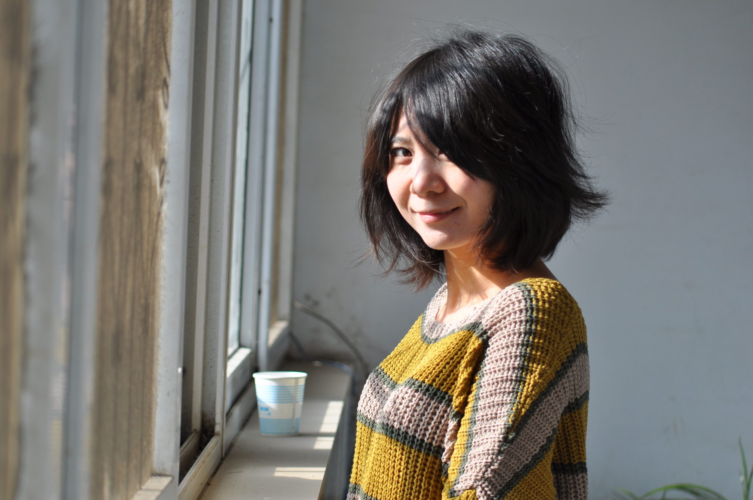 A young woman with a bob hairstyle smiles warmly while standing by a window indoors.
